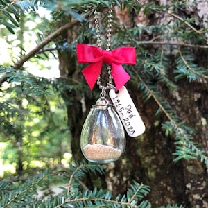 May include: A glass ornament with sand inside, hanging from a red bow and a silver chain. A silver tag with the text "Dad 1965-2020" is attached.