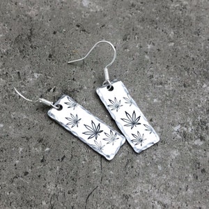 Peut inclure: Une paire de boucles d'oreilles pendantes en argent avec un motif rectangulaire orné d'un motif de feuilles de marijuana noires.