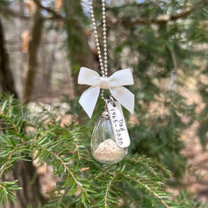 May include: A silver chain necklace with a white satin bow and a glass teardrop pendant filled with sand. The pendant is attached to a silver tag with the inscription "Dad 1965-2020".
