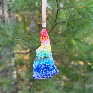 May include: A colorful ornament in the shape of the state of New Hampshire, with a rainbow gradient of colors. The ornament is suspended by a light pink ribbon, perfect for holiday decor.
