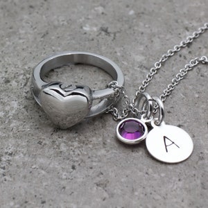 Op de afbeelding: Een zilveren ring met een hartvormige hanger en een zilveren ketting met een paarse kristallen hanger en een zilveren hanger met de letter 'A' erop.