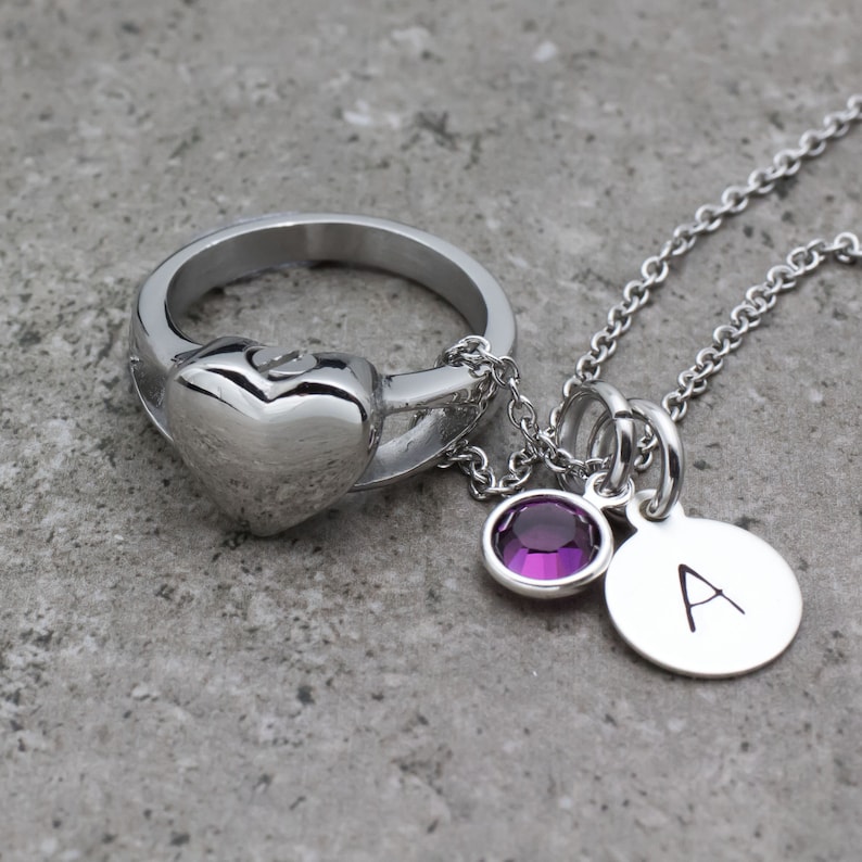 Op de afbeelding: Een zilveren ring met een hartvormige hanger en een zilveren ketting met een paarse kristallen hanger en een zilveren hanger met de letter 'A' erop.