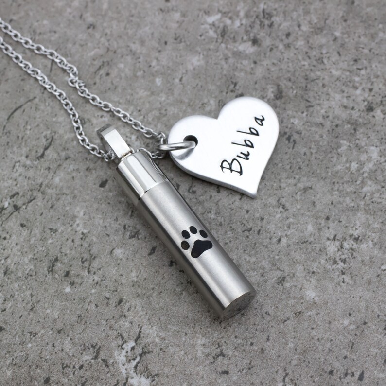 May include: Silver necklace with a heart charm engraved with the name "Bubba" and a cylindrical cremation urn pendant with a paw print.