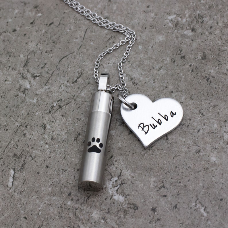 May include: Silver necklace with a small cylinder charm engraved with a paw print and a heart charm engraved with the name "Bubba".