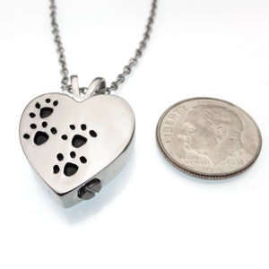Puede incluir: Collar de cremaci&oacute;n de coraz&oacute;n plateado con cierre de tornillo y recortes de huellas de patas. El collar se muestra junto a una moneda de diez centavos de d&oacute;lar estadounidense para comparar el tama&ntilde;o.