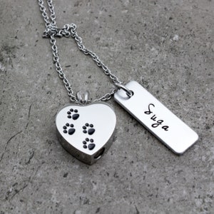 Puede incluir: Collar de plata con un colgante en forma de coraz&oacute;n con huellas de patas y una etiqueta de plata grabada con el nombre "SUGA".