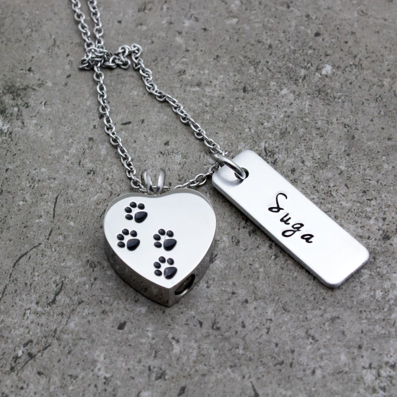 Puede incluir: Collar de plata con un colgante en forma de coraz&oacute;n con huellas de patas y una etiqueta de plata grabada con el nombre "SUGA".