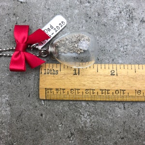 May include: A silver necklace with a red bow and a glass vial containing a small amount of white sand. The vial is attached to a silver tag that reads "Dad 1968-2020".