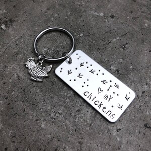 K&ouml;nnte beinhalten: Silberner Schl&uuml;sselanh&auml;nger mit einem kleinen H&uuml;hneranh&auml;nger und einem rechteckigen Anh&auml;nger mit der Aufschrift "Ich liebe meine H&uuml;hner" mit einem Herzsymbol und H&uuml;hnerpfoten um den Text herum.