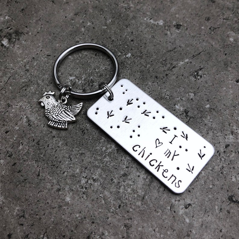 K&ouml;nnte beinhalten: Silberner Schl&uuml;sselanh&auml;nger mit einem kleinen H&uuml;hneranh&auml;nger und einem rechteckigen Anh&auml;nger mit der Aufschrift "Ich liebe meine H&uuml;hner" mit einem Herzsymbol und H&uuml;hnerpfoten um den Text herum.