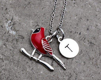 May include: A silver necklace featuring a red cardinal bird pendant perched on a branch. The bird has black accents and is paired with a round charm engraved with the letter "T". The necklace is on a gray surface.