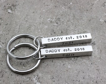 May include: Silver keychain with two bar-shaped charms. The charms are engraved with "DADDY est. 2018" and the names "Jude" and "Eddie".