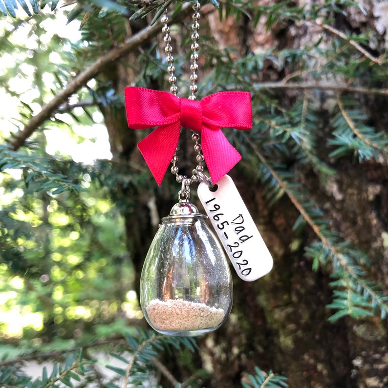 May include: A clear glass teardrop-shaped ornament filled with sand, hanging from a silver chain with a red bow. A silver tag with the text "Dad 1965-2020" is attached to the chain.