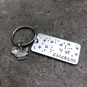 K&ouml;nnte beinhalten: Silberner Schl&uuml;sselanh&auml;nger mit einem kleinen H&uuml;hneranh&auml;nger und einem rechteckigen Anh&auml;nger mit der Aufschrift "Ich liebe meine H&uuml;hner" in Braille.