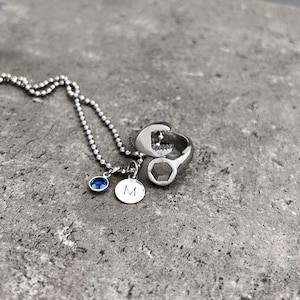 May include: A silver necklace with a blue gemstone and a small round charm with the letter "M". A silver ring shaped like a wrench is also present. The jewelry is displayed on a gray stone surface.