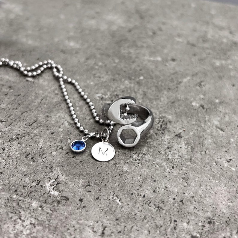 May include: A silver necklace with a blue gemstone and a small round charm with the letter "M". A silver ring shaped like a wrench is also present. The jewelry is displayed on a gray stone surface.