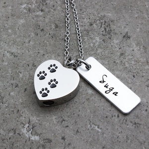 Puede incluir: Collar de cremaci&oacute;n de coraz&oacute;n plateado con huellas de patas y una etiqueta plateada grabada con el nombre "SUGA"