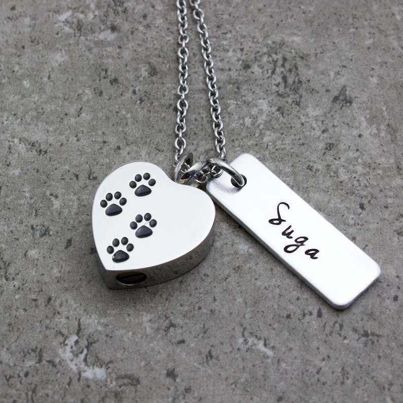 Puede incluir: Collar de cremaci&oacute;n de coraz&oacute;n plateado con huellas de patas y una etiqueta plateada grabada con el nombre "SUGA"