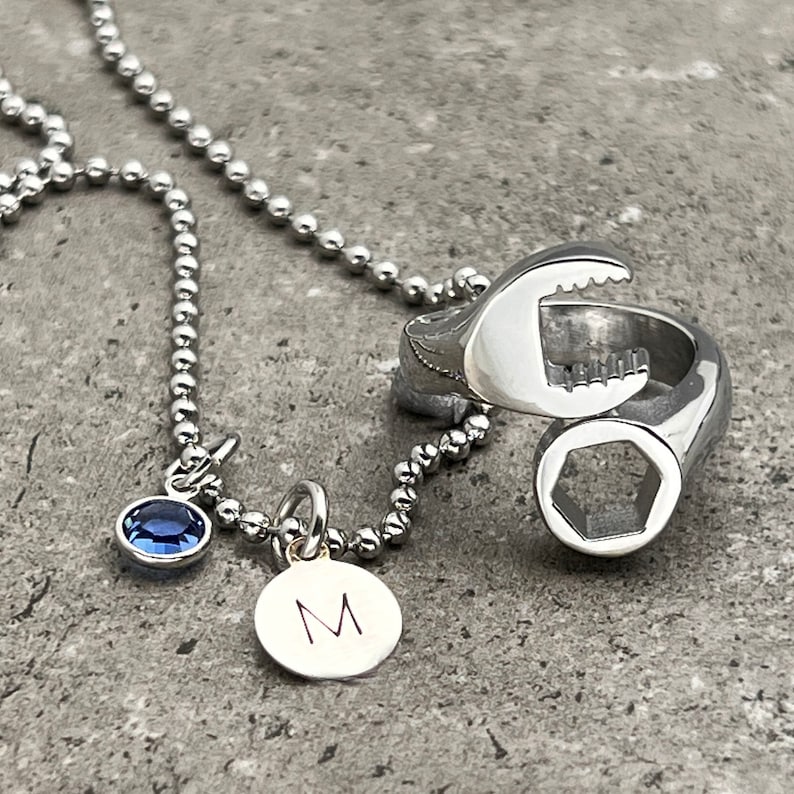 May include: A silver necklace featuring a wrench pendant, a blue gemstone, and a small circular charm with the letter "M". The necklace has a ball chain design. The wrench pendant is a ring.