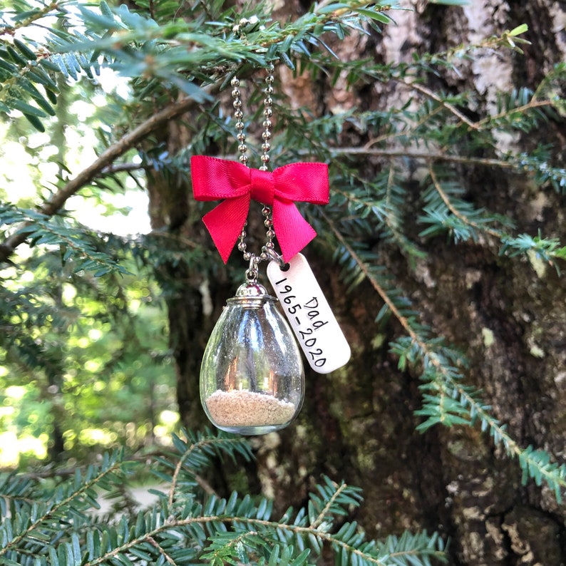 May include: A glass ornament with sand inside, hanging from a red bow and a silver chain. A silver tag with the text "Dad 1965-2020" is attached.