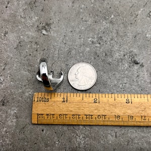 May include: A silver-colored ring with an anchor design, next to a US quarter and a wooden ruler. The ring is approximately 1 inch in length. The background is a mottled gray surface.