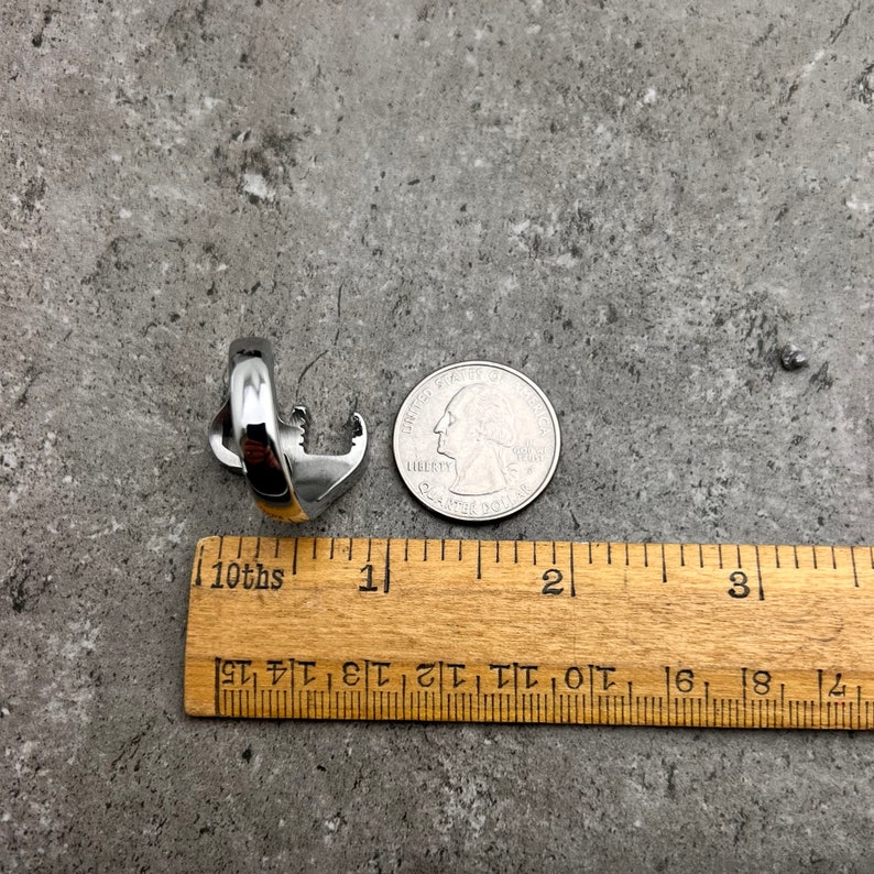 May include: A silver-colored ring with an anchor design, next to a US quarter and a wooden ruler. The ring is approximately 1 inch in length. The background is a mottled gray surface.