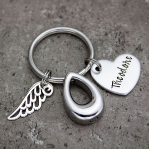 May include: Silver keychain with a heart charm engraved with the name "Theodore", a teardrop-shaped cremation urn charm, and a small angel wing charm.