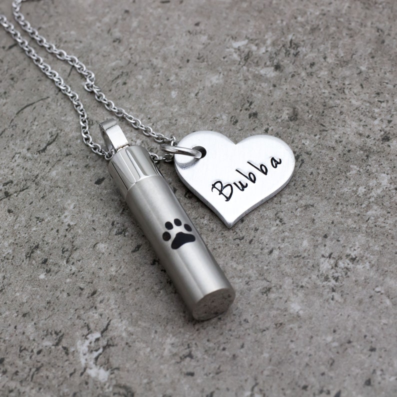 May include: Silver necklace with a small cylinder pendant featuring a paw print and a heart-shaped pendant with the name "Bubba" engraved on it.