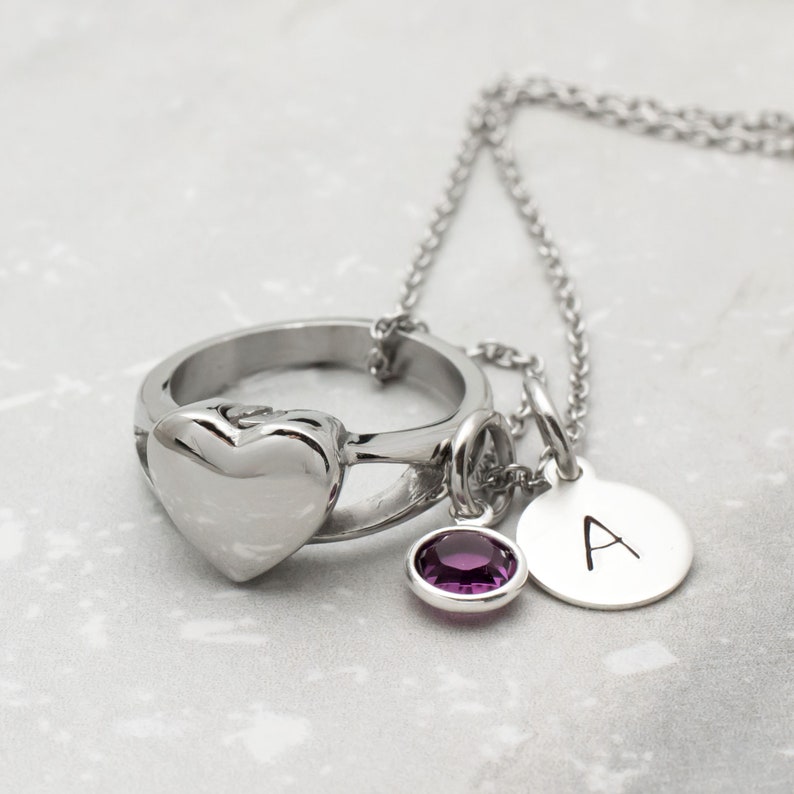 Op de afbeelding: Een zilveren ketting met een hartvormige ringbedel, een kleine paarse edelsteenbedel en een ronde bedel met de letter 'A' erop gegraveerd.