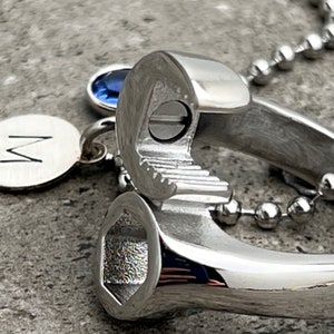 May include: A silver wrench pendant with a small blue gemstone and a round charm with the letter "M". The pendant is attached to a ball chain necklace. The wrench has a detailed, realistic design.