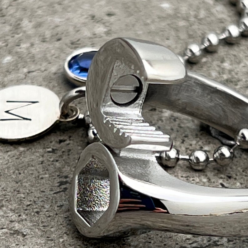 May include: A silver wrench pendant with a small blue gemstone and a round charm with the letter "M". The pendant is attached to a ball chain necklace. The wrench has a detailed, realistic design.