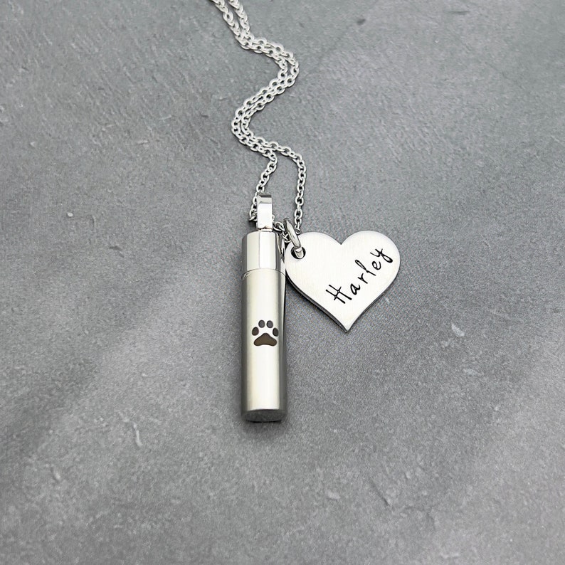 May include: Silver necklace with a small cylindrical urn engraved with a paw print and a heart-shaped pendant engraved with the name "Harley".