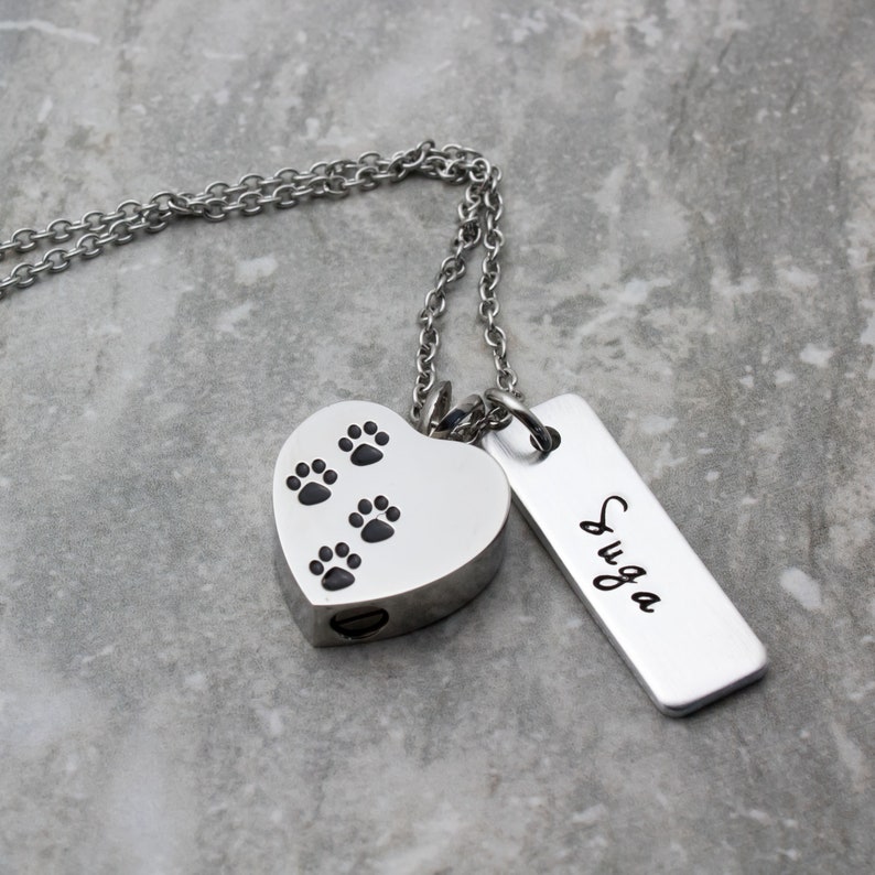 Puede incluir: Collar de plata con un colgante en forma de coraz&oacute;n con huellas de patas y una peque&ntilde;a etiqueta rectangular grabada con el nombre "SUGA".