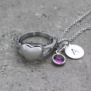 Op de afbeelding: Een zilveren ring in de vorm van een hart met een kleine paarse edelsteenbedel en een zilveren bedel met de letter 'A' aan een zilveren ketting.