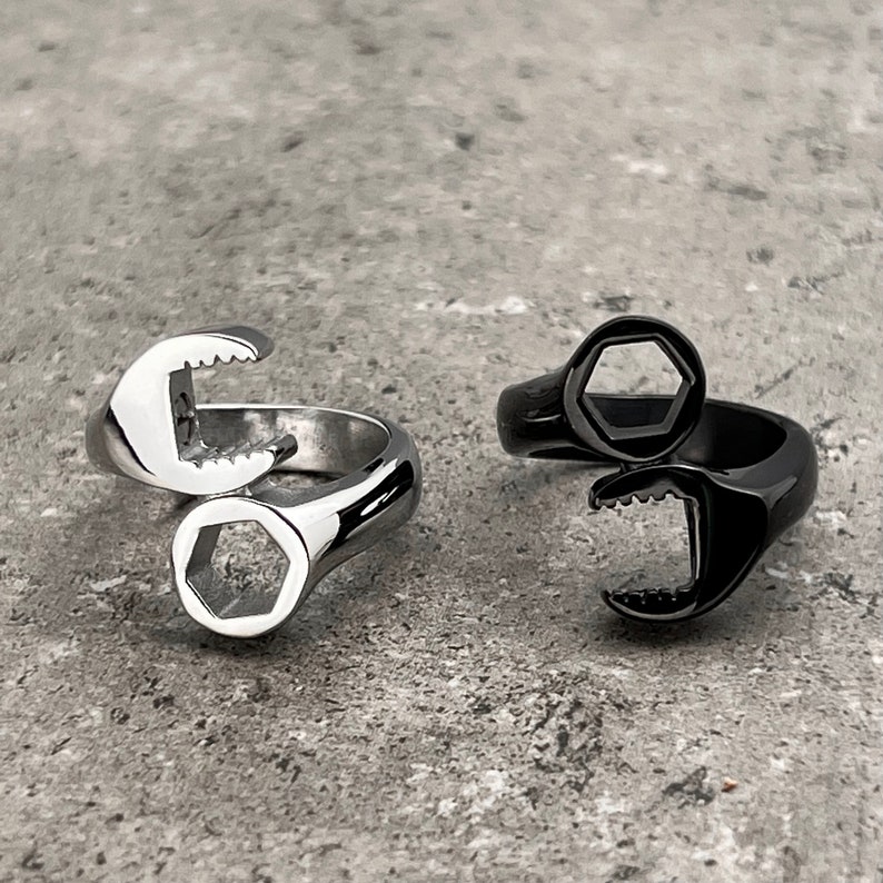 May include: Two rings with a wrench design. One ring is silver, and the other is black. Each ring features a wrench head and a hexagonal opening. The rings are set against a gray background.