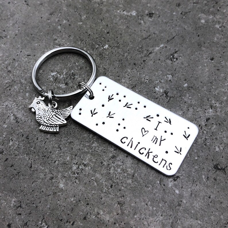 K&ouml;nnte beinhalten: Silberner Schl&uuml;sselanh&auml;nger mit einem kleinen H&uuml;hneranh&auml;nger und einem rechteckigen Anh&auml;nger mit der Aufschrift "Ich liebe meine H&uuml;hner" mit einem Herzsymbol und einem Muster aus H&uuml;hnerpfoten.