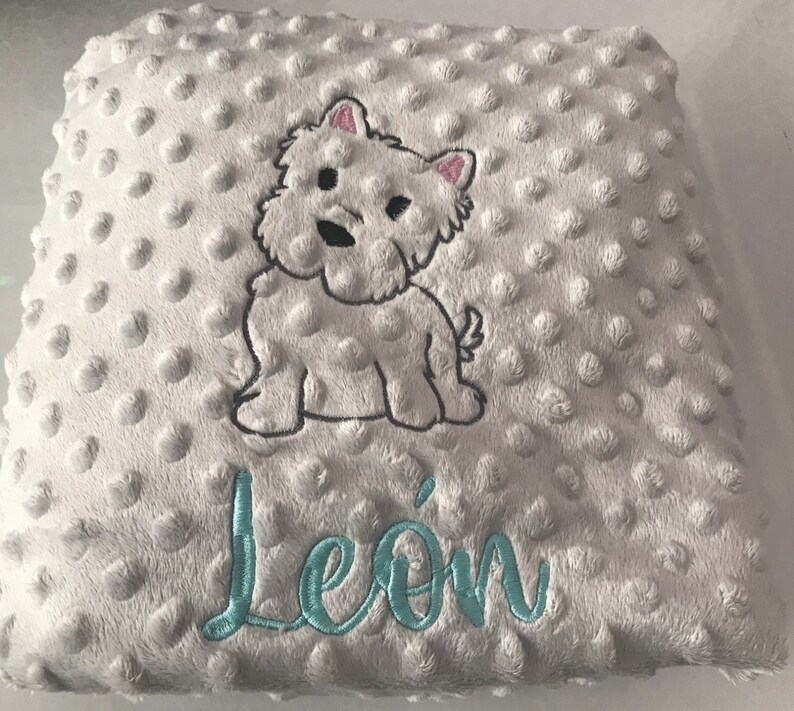 Op de afbeelding: Een wit pluche babydeken met een witte geborduurde hond en de naam "Le&oacute;n" in tealblauw.