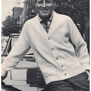 May include: A man wearing a white cardigan sweater with a button closure, standing next to a black car. He is smiling and looking at the camera.