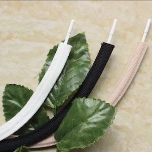 May include: Three shoelaces in different colors: white, black, and beige. The shoelaces are made of a soft, stretchy material and have a flat, rounded shape.