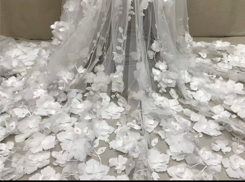 May include: White floral lace fabric with 3D flowers and pearl embellishments. The fabric is sheer and delicate, perfect for special occasion garments.