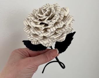 Puede incluir: Una sola rosa blanca de papel hecha con las páginas de un libro. La rosa tiene un tallo y hojas negras.