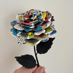 Puede incluir: Una rosa de c&oacute;mic colorida con hojas y tallo de papel negro. La rosa est&aacute; hecha de p&aacute;ginas de un c&oacute;mic con texto e im&aacute;genes. El texto en la rosa incluye "GOODNS! 0009" y "1,EK, KNO".