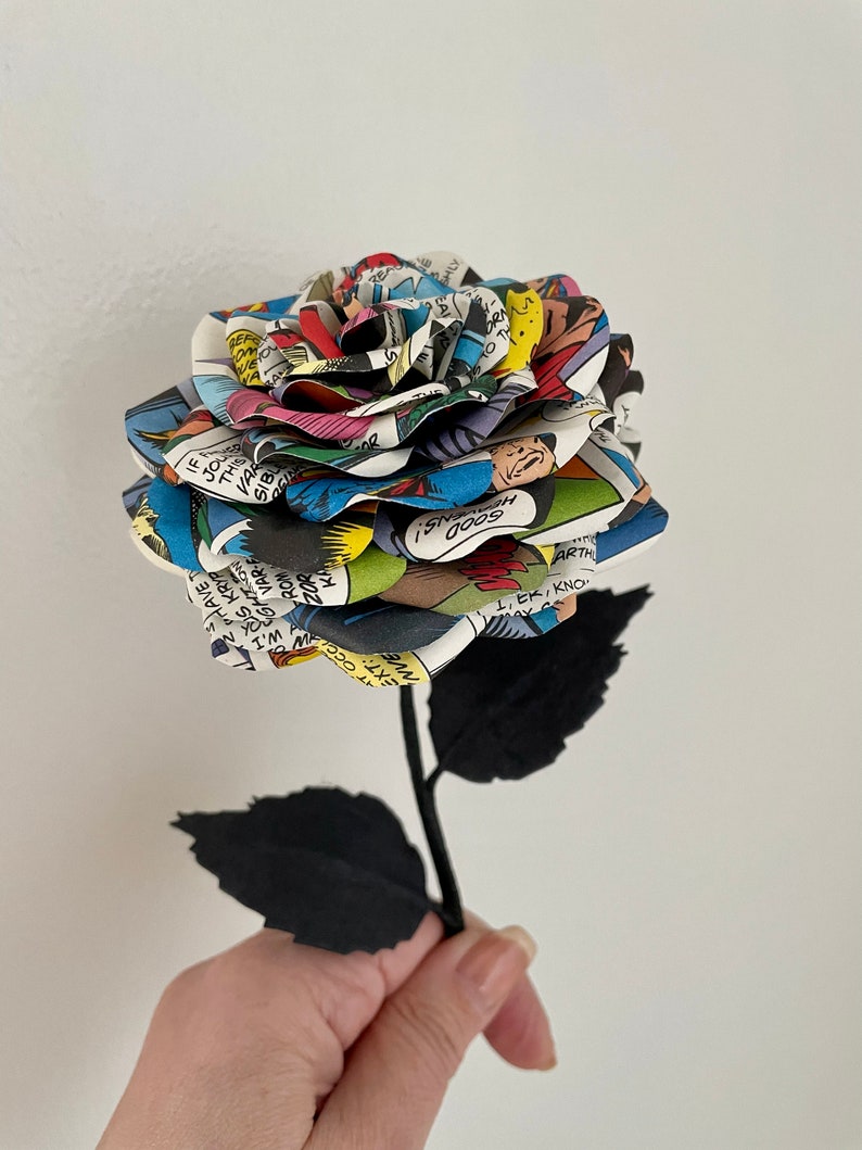 Puede incluir: Una rosa de c&oacute;mic colorida con hojas y tallo de papel negro. La rosa est&aacute; hecha de p&aacute;ginas de un c&oacute;mic con texto e im&aacute;genes. El texto en la rosa incluye "GOODNS! 0009" y "1,EK, KNO".