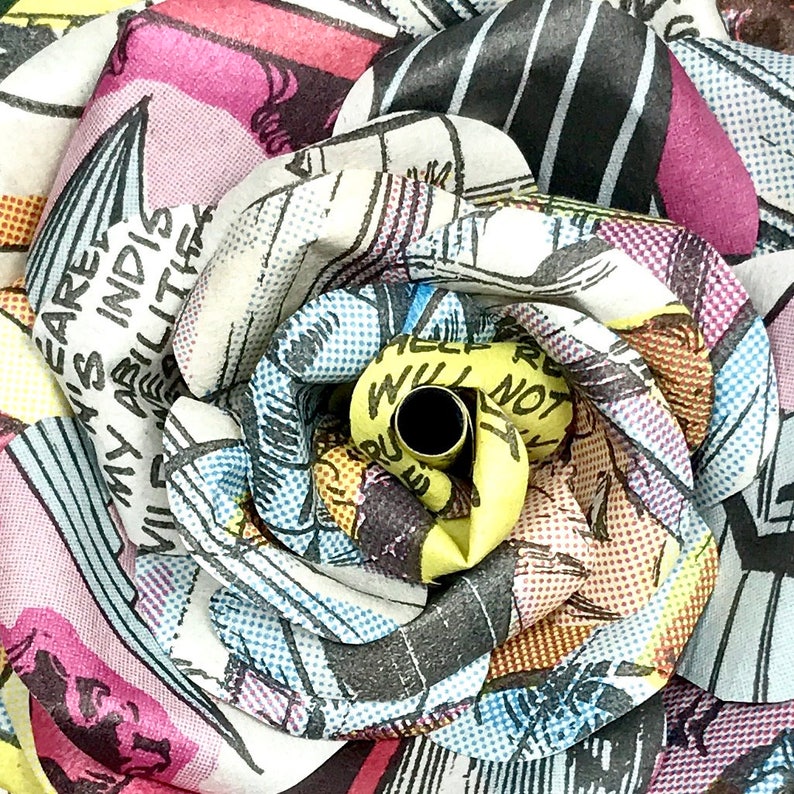 May include: A close-up of a multi-colored fabric rose made from comic book pages. The rose is yellow in the center and has a black hole in the center. The rose is surrounded by petals of different colors, including pink, blue, and yellow.