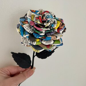 Puede incluir: Un solo tallo negro con hojas negras sostiene una rosa hecha de p&aacute;ginas de c&oacute;mics. La rosa es multicolor y presenta una variedad de personajes y textos de c&oacute;mics. El texto incluye "IF YOUR THIS VAR SIBL REF WEION A HAVE T S KRYP VAR ROM KA 200 EXTOCCAW W.I VENS! 0009 ARTH KNO OP CO READKIE B SHLY REA EAL ORMA TO TH"