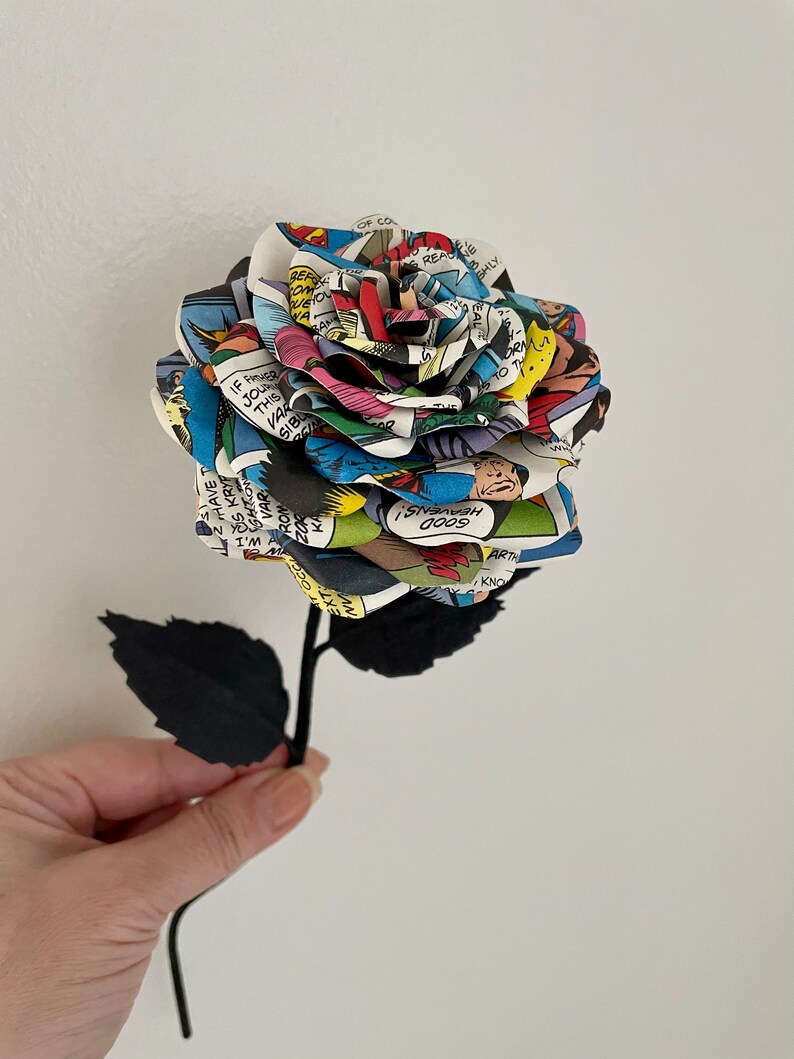 Puede incluir: Un solo tallo negro con hojas negras sostiene una rosa hecha de p&aacute;ginas de c&oacute;mics. La rosa es multicolor y presenta una variedad de personajes y textos de c&oacute;mics. El texto incluye "IF YOUR THIS VAR SIBL REF WEION A HAVE T S KRYP VAR ROM KA 200 EXTOCCAW W.I VENS! 0009 ARTH KNO OP CO READKIE B SHLY REA EAL ORMA TO TH"