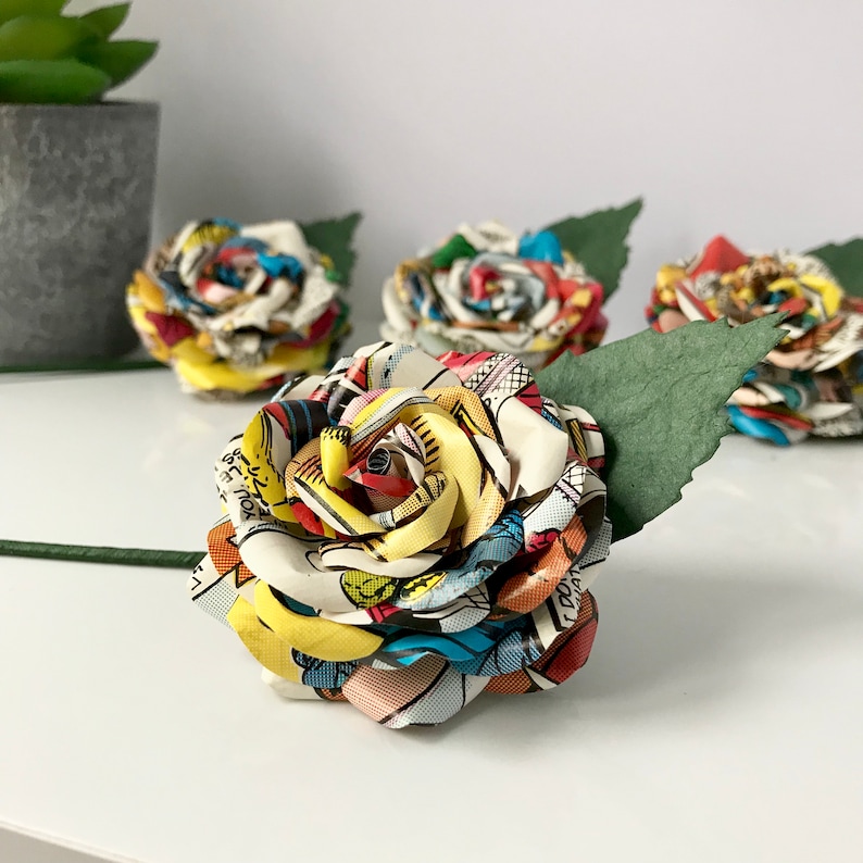 May include: Four colorful paper roses made from comic book pages. The roses are arranged in a row with a green leaf stem.