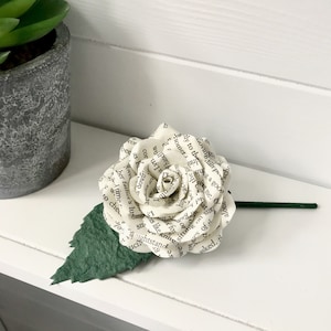 May include: A white paper rose crafted from the pages of a book. The rose features a green stem and leaf. This rose is a unique and beautiful way to express love or appreciation.