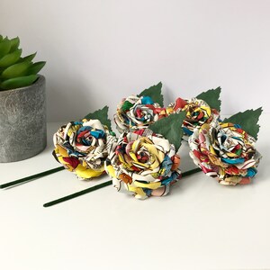 May include: Five comic book paper roses with green stems. The roses are made from colorful comic book pages and are arranged in a bouquet.