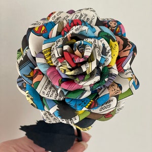 Puede incluir: Una rosa de papel hecha de p&aacute;ginas de c&oacute;mics con Superman, Batman y otros superh&eacute;roes. La rosa es una mezcla vibrante de rojo, azul, amarillo y verde. El tallo es negro.
